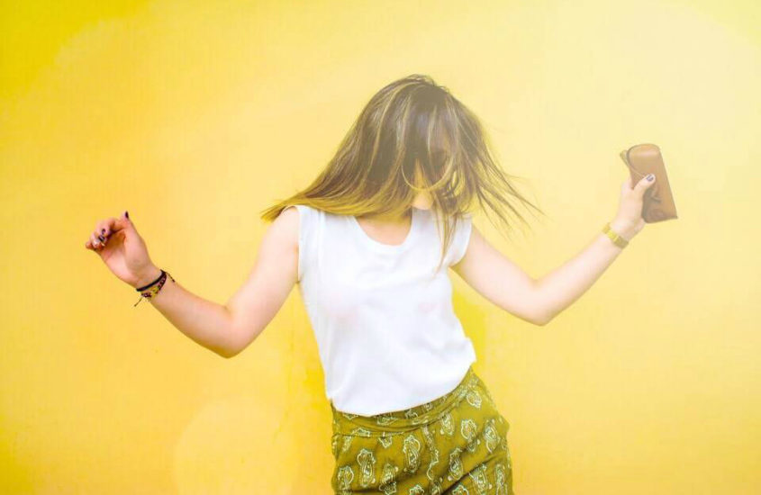 Blonde woman standing against a yellow wall, with her arms out to either side either dancing or celebrating, with her hair covering her face