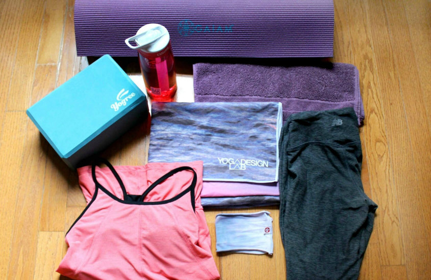 Yoga gear laid out on a wood floor including a yoga mat, yoga block, water bottle, yoga towel, towel, leggings, work out top, and headband