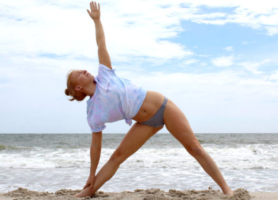 Redhead woman standing on the beach on the sand in front of the ocean doing triangle yoga pose