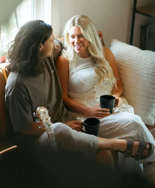 A couple sitting on a couch sipping cups of coffee, turned toward eachother talking