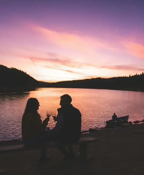 A couple with glasses of wine in their hands with a sunset behind them on a lake