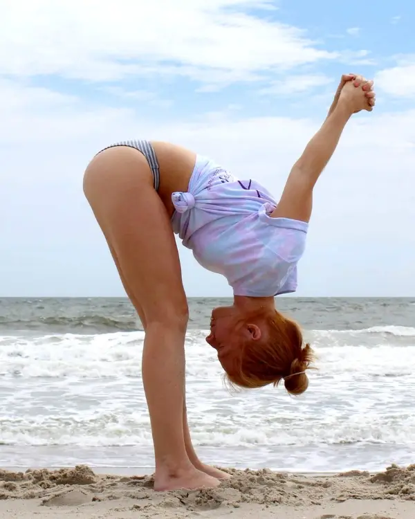 Small redhead woman doing forward fold with arms behind variation yoga pose on the beach in front of the ocean