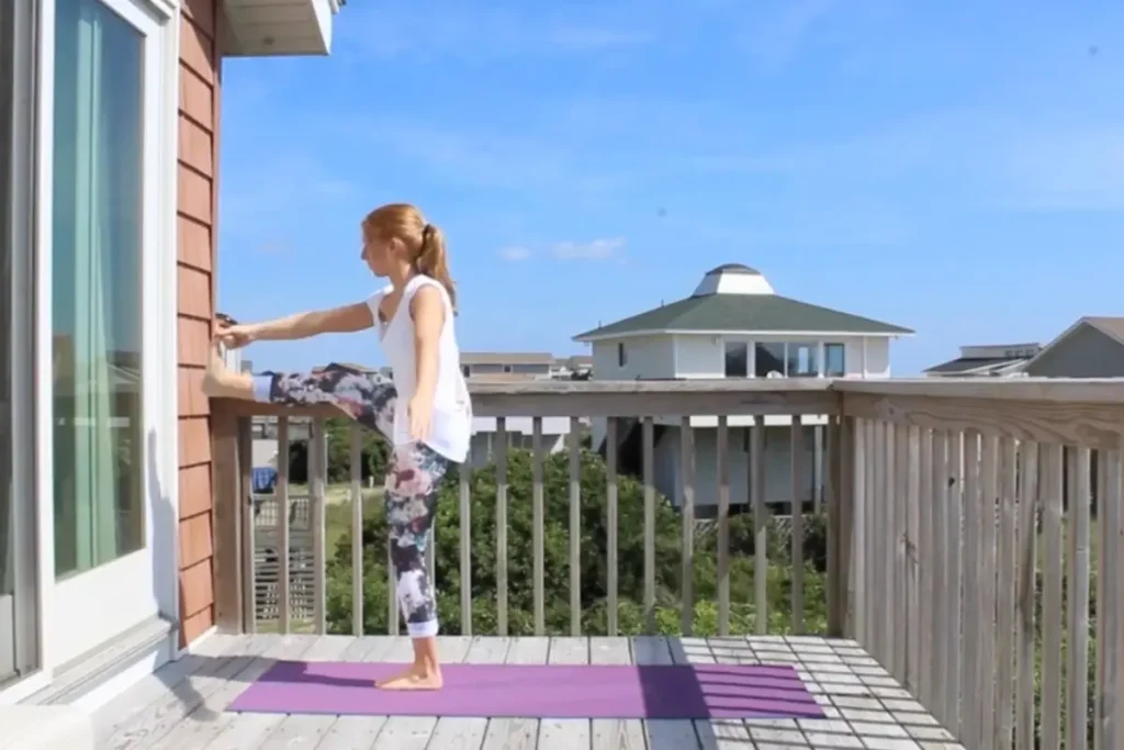 Redhead woman doing the hand to big toe yoga post, leg in front of her, on a sunny deck