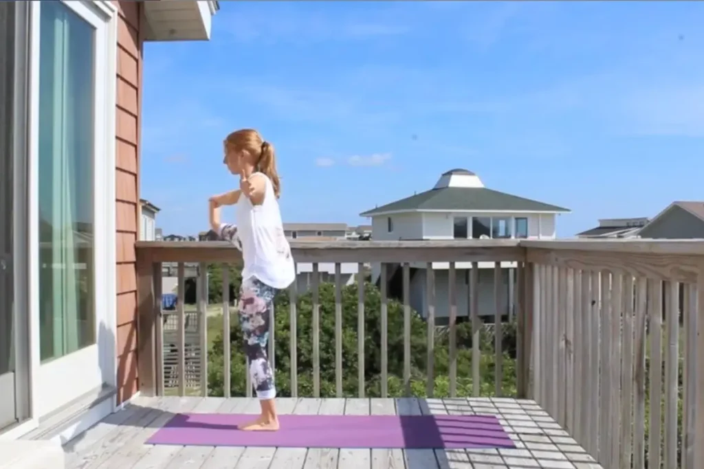 Redhead woman doing the hand to big toe yoga post, leg to the side of her, on a sunny deck