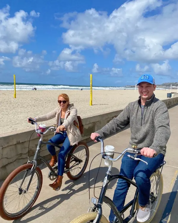 A happy couple biking down the boardwalk with the beach in the background on a sunny day