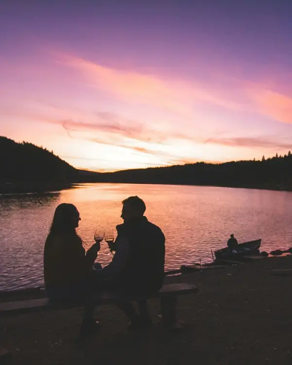 A couple with glasses of wine in their hands with a sunset behind them on a lake
