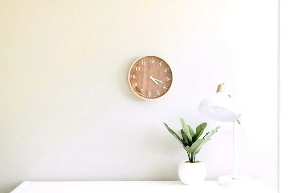 simple living desk room Simple desk set up with a wooden clock, small white vase with a green fern, and white lamp against a yellow wall