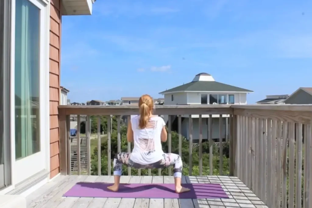 Redhead woman doing the buddha squats yoga pose, squatting low, on a sunny deck
